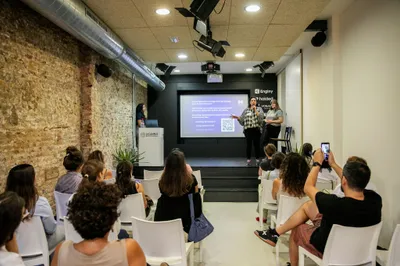 The full room watching two speakers on stage, the partner logos on the wall beside the screen.